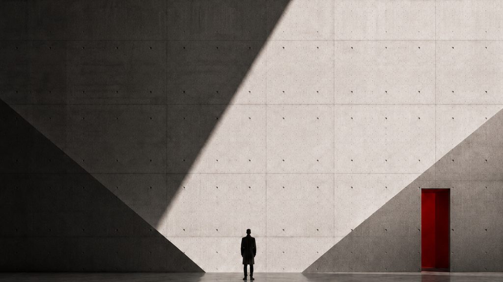 A lone figure in a vast concrete hall, a hard diagonal of daylight across the wall and one red doorway beyond.